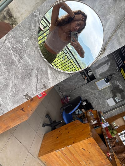 This image captures a woman's selfie in a mirror, against the backdrop of what appears to be a shop interior, likely located in Quản Bạ Commune, Vietnam. The woman is holding her phone, posing in front of a round mirror. The reflection shows her wearing a tank top and green shorts, with a view of mountainous terrain and green vegetation outside.
The shop interior features a grey marble-like countertop where the mirror is placed. In the background, there is a wooden counter with items like coconuts and other goods on display. A blue chair is situated near the counter. The walls are a grey textured material, adorned with a wall-mounted fan, a framed document, and a menu board with Vietnamese text and prices. The floor is tiled, and there is a wooden structure beside the countertop. A sticker with a QR code is visible on the wooden structure.