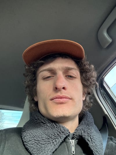 A male individual is depicted in a close-up, head-and-shoulders, selfie-style photograph taken inside a vehicle. The subject, a male with fair skin and dark, curly hair, wears an orange baseball cap and a dark jacket featuring a prominent black textured collar and a silver zipper. His eyes are directed towards the camera, maintaining a neutral expression with closed lips. A seatbelt diagonally crosses his right shoulder and chest. The interior of the vehicle is visible, including a light-colored headliner, a grey plastic grab handle on the upper right, and a light-colored seat behind him. Blurred outdoor light and faint environmental details are visible through the vehicle's windows on both sides. The scene is located within a vehicle in the city of Fontenay-sous-Bois, France.
