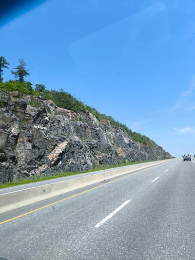 An outdoor, eye-level, wide shot shows a highway next to a rocky cliff face on a clear, sunny day. The road, paved with asphalt, is divided into two lanes by white lines, with a yellow line marking the edge closest to the viewer. A concrete barrier separates the road from the cliff, which is covered in rocks and sparse green vegetation. The cliff is rugged, with varying shades of gray and brown rock formations and patches of pinkish-orange mineral deposits. A few trees with green foliage grow at the top of the cliff. In the distance, a white pickup truck drives on the right lane of the highway. The sky is a bright, clear blue with a few wispy white clouds. The scene appears to be a scenic view of a highway in a natural setting.