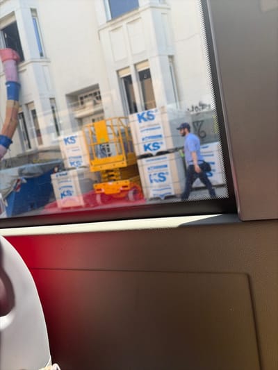 This image captures a reflection in a window, likely from a vehicle, offering a glimpse of an urban scene in Münster, Germany. In the background, a modern white building with numerous windows and balconies is visible. In the foreground, a construction site is suggested by stacks of white boxes with blue lettering that appears to be "KS" and a yellow scissor lift. A man wearing a blue shirt, dark pants, and a baseball cap is walking past the construction materials. The lower part of the image shows the interior of the vehicle, with a textured surface and a red illuminated area. The scene is illuminated by sunlight, casting shadows and creating a somewhat blurry, reflective quality to the overall image.