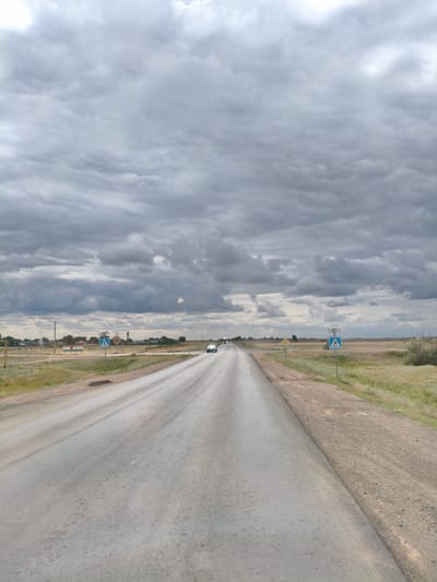 The image shows a straight asphalt road stretching into the distance under a cloudy sky. The road is flanked by dry grass and sparse vegetation. On the right side of the road, there is a pedestrian crossing sign and a yellow diamond-shaped warning sign. In the distance, there are some buildings and trees, indicating a settlement. A white car is driving on the road away from the camera. The overall environment appears rural and somewhat desolate. The location is identified as Бударин ауылдық округі, Kazakhstan.