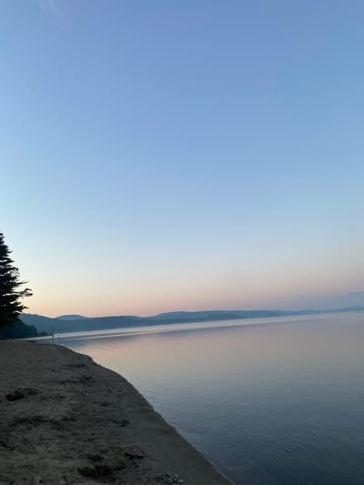 The image depicts a serene lake scene at dawn or dusk, with a sandy shoreline in the foreground. A large evergreen tree stands on the left side of the beach. The water is calm, reflecting the soft hues of the sky, which transitions from a pale blue at the top to shades of pink and purple near the horizon. Distant hills, covered in a light mist, form a hazy silhouette across the landscape. Two wooden posts are visible on the sandy beach, one closer to the water and another further back. The environment appears tranquil and natural, with no signs of human activity or urban presence. The image is taken from a slightly elevated perspective, looking out over the lake.
