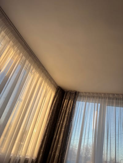 The image captures an interior scene within a room in Tsenovo, Bulgaria, viewed from a low angle looking upwards towards a corner. The upper right portion of the frame features a plain, light-colored ceiling. Below the ceiling, two window sections are covered by curtains. On the left, light-toned, sheer curtains are drawn, allowing warm, golden sunlight to intensely illuminate them and cast a glow onto the adjacent ceiling. A white curtain track is visible above these curtains. To the right, darker, patterned curtains are partially drawn, revealing a second set of sheer, light-toned curtains behind them. Through these sheer curtains, a view of the exterior is visible, showing a clear blue sky and bare-branched trees, consistent with daytime conditions. No individuals, actions, or interactions are present.