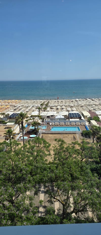 This aerial shot shows a beach in Riccione, Italy. The beach is filled with rows of beach umbrellas and sunbeds, mostly yellow, extending along the shoreline. The sea is calm and blue. Adjacent to the beach is a complex with pools, including a long rectangular pool and a smaller, circular pool. Trees are growing next to a walkway.