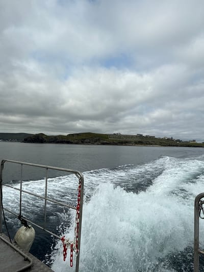 The image depicts a view from a moving boat in the waters off the coast of Lerwick, United Kingdom. The boat is traveling away from a landmass that features rolling green hills dotted with buildings and what appears to be a graveyard on a ridge. The water behind the boat is turbulent, creating a large wake with white foam. A metal railing runs along the edge of the boat, with a white buoy and a red and white chain attached to it. The sky is overcast with thick grey clouds, with patches of blue sky visible. The overall scene conveys a sense of travel and exploration in a coastal environment.