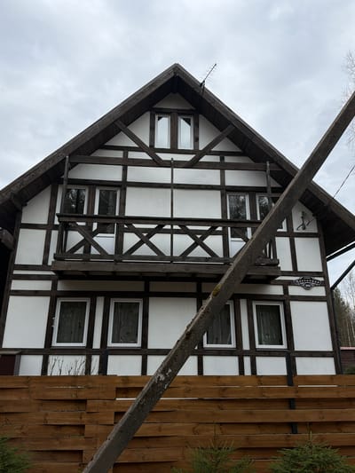 The image shows a two-story building with a distinctive half-timbered facade, characterized by dark brown wooden beams forming a grid pattern on a white background. The building has a steeply pitched roof with a sharp gable end visible on the left. A sign with Russian text and the number 982 is affixed to the upper part of the facade. Several windows with white frames are visible. A wooden fence runs along the right side of the building. A diagonal wooden beam obscures part of the view. The location appears to be Glubokoe, Russia, based on the signage. The sky is overcast.