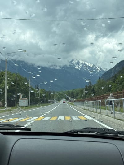 The image, taken from inside a vehicle, shows a scenic road in Верхняя Теберда, Russia, leading towards snow-capped mountains under a cloudy sky. The road features a pedestrian crossing with yellow and white stripes. On the right side of the road, a person wearing a hooded jacket stands near a metal fence and a wooden fence. Streetlights line the left side of the road, adjacent to trees and a small utility building. Water droplets are visible on the car's windshield.