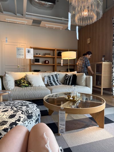 This image depicts an interior scene at an IKEA store in Goyang-si, South Korea. In the foreground, a portion of a person's legs is visible, seated on a circular, patterned ottoman. The ottoman features a black and white floral design. In the mid-ground, a light-colored sofa with several decorative pillows is arranged, including one with a green and black abstract pattern and another with a black and white geometric design. A round glass-topped coffee table with a light wood base sits in front of the sofa, holding a tray with two gold candle holders and a glass beaker. The background reveals a wooden shelving unit filled with various items, including books and decorative objects. A floor lamp with a light-colored shade stands to the left of the shelving unit. A woman in a plaid shirt is visible in the right mid-ground, opening a cabinet with woven rattan detailing. A clock is mounted on the wood-paneled wall behind her. The ceiling features a modern chandelier made of glass tubes. Several IKEA price tags and informational posters are hanging from the ceiling and attached to furniture. The floor is covered with a patterned rug in shades of gray, beige, and brown.