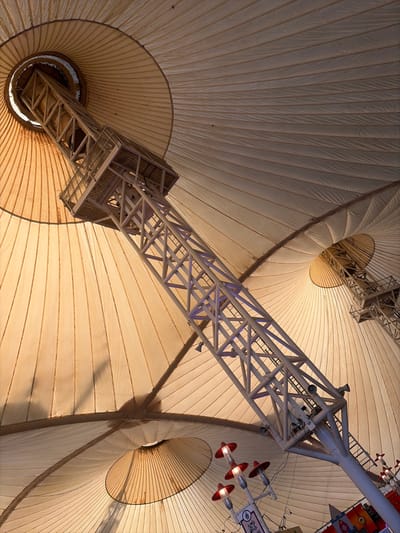 The image displays the interior of the Skyline Pavilion at Butlin's Skegness, located in the city of East Lindsey, United Kingdom. The scene is dominated by a vast, cream-colored tensile fabric roof, characterized by radial pleats and multiple conical structures where the fabric is tensioned upwards. A large, white, open-lattice metal support tower extends diagonally from the lower right to the upper left of the frame. This tower features a small observation or equipment platform near its upper end and has several dark-colored speakers or lights mounted along its length, some illuminated with a subtle purple light. A vertical ladder is visible on the right side of the main support tower. In the lower right quadrant, a lamppost-style fixture with three downward-facing, red-hooded lights is present, partially obscuring a white sign with blue text. The lighting suggests an indoor environment, with natural light potentially filtering through the fabric and augmented by artificial illumination, creating variations in shadow and light across the ceiling. No individuals are visible.