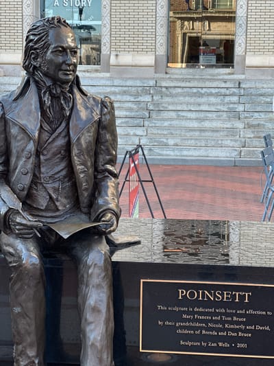 A bronze statue of Joel Roberts Poinsett sits in front of a building. The statue depicts a seated man in a coat, holding a paper. Behind the statue is a black plaque that reads "POINSETT" and dedicates the sculpture to Mary Frances and Tom Bruce by their grandchildren. It identifies Zan Wells as the sculptor and dates the work to 2001. A sign with the words "A STORY" is visible in the reflection of a window in the background.