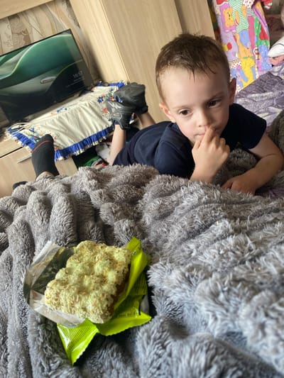 The image shows a young boy lying on a fuzzy gray blanket. He is wearing a dark blue t-shirt and black socks. His right hand is touching his chin, and he appears to be looking off to the side. In the foreground, there is a snack, possibly a cookie or bar, wrapped in green and silver packaging. The background is slightly blurred but shows a television screen displaying a nature scene with a time of 16:57. There are also some colorful blankets and a wooden wardrobe or furniture visible. The scene appears to be in a bedroom or living room. The location is identified as Ihnatpil, Ukraine.