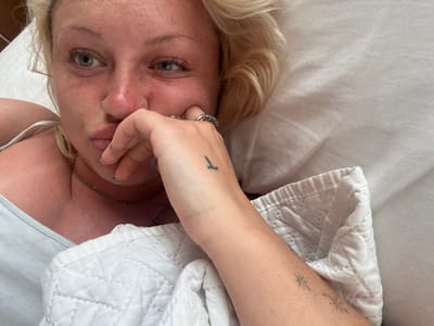 A female individual is depicted in a close-up, lying in what appears to be a bed, located in Olde Yard Village, British Virgin Islands. She possesses blonde hair, fair skin, and light-colored eyes that exhibit slight redness. Her facial expression includes a pouted mouth, and her gaze is directed towards the upper left of the frame. She is wearing a light-colored tank top or camisole. Her right hand is raised, with fingers partially covering her mouth and nose. This hand displays multiple rings on the fingers and a small black, vertical line tattoo on its back. Her fingernails are painted with a dark, patterned polish. Two circular nose piercings are present. A thin gold-colored necklace is visible around her neck. Her right forearm, partially exposed, features two distinct star-like tattoos. She is partially covered by a white quilted blanket or duvet, resting against a white pillow. The immediate environment consists of white bedding, indicative of an indoor setting.