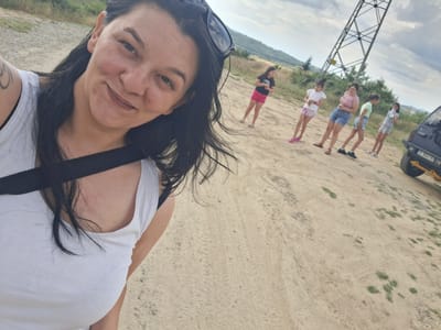 A woman is taking a selfie outdoors with a group of people in the background. She is in the foreground, wearing a white tank top and has black hair. She is smiling and looking at the camera. In the background, there are several other people, including younger girls, standing on a dirt path. Behind them, there is a large metal tower, hills covered in vegetation, and a cloudy sky. A vehicle is partially visible on the right side of the image. The setting appears to be a rural or outdoor area, possibly in Dobrotino, Bulgaria, given the provided location context. The weather seems overcast. The overall mood seems casual and familial, as if the group is on an outing. No discernible text is visible in the image.