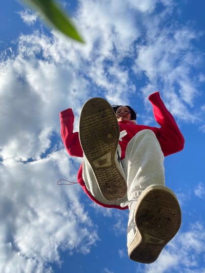 A low-angle shot shows a person standing over the camera, set against a sky with scattered clouds. The person wears a red sweatshirt and white pants, with worn white sneakers. The soles of the sneakers are prominently displayed. A blurred green object is visible in the lower left corner of the frame. The location, Doma 45 Kilometr, Russia, is not discernible from the image.