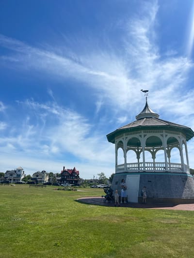 The image shows a gazebo with a whale weather vane on top, set against a bright blue sky with wispy clouds. The gazebo is white with green accents and features an open, colonnaded design. In front of the gazebo, several people are gathered on a paved area, including a woman with a baby stroller and two children. Behind the gazebo, a row of Victorian-style houses with red roofs can be seen, characteristic of the Oak Bluffs area. The scene appears to be a sunny day, likely in the late morning or early afternoon, given the bright light and clear sky. The overall atmosphere is relaxed and summery, with the people enjoying the outdoors and the picturesque setting.