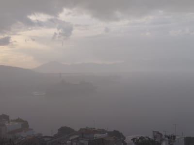The image depicts a hazy, overcast day over the Municipal Unit of Aegio, Greece. In the foreground, residential buildings with red-tiled roofs are visible. Beyond the buildings, a body of water, likely the Gulf of Corinth, is obscured by mist. A large ship, possibly a cruise liner, is visible in the distance on the water. Mountains form a faint outline on the horizon, further softened by the atmospheric conditions. The overall impression is one of diffused light and limited visibility, suggesting a misty or foggy atmosphere.