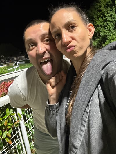 A man and woman are posing for a selfie outdoors at night in the Borough of Spelthorne, United Kingdom. The man, who is behind the woman, is wearing a light grey t-shirt and sticking his tongue out. The woman is wearing a grey jacket. There are plants and a metal fence visible in the background.