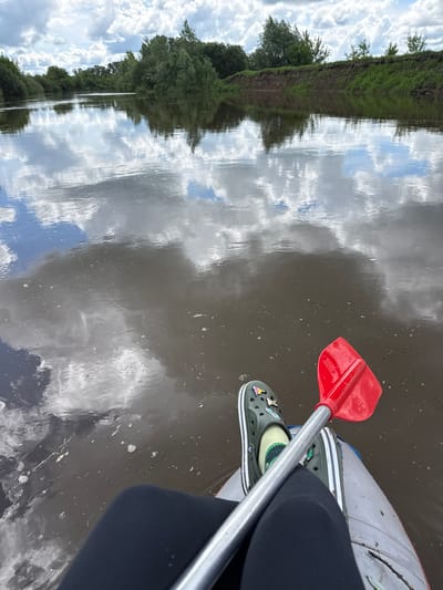 The image shows a person kayaking on a body of water in Izh-Boyki, Russia. The kayak is grey and white, with a person wearing black leggings and green Crocs inside. The kayak paddle is red and silver and lies across the person's lap. The water reflects the sky, which is filled with clouds. Trees and greenery line the water's edge on one side.
