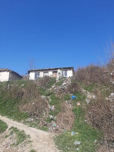 The scene, located in Ivanski, Bulgaria, depicts a rural or semi-rural landscape under a clear blue sky. In the foreground, an unpaved dirt path, light brown in color, meanders uphill from the lower left towards the center. The path is bordered by low-lying green vegetation and patches of dry, brown, bushy plants. The immediate surroundings of the path and the ascending hillside are heavily strewn with various types of refuse, including plastic bottles, white plastic bags, and multiple prominent blue plastic bags.
The midground is dominated by a steep, overgrown hillside covered with a mix of green ground cover and significant clusters of dry, reddish-brown thorny or bushy vegetation. This entire slope exhibits a substantial accumulation of discarded items and debris.
In the background, positioned at the crest of the hill, are two dilapidated single-story structures. The more prominent building, slightly right of center, features light-colored, possibly whitewashed, walls displaying cracks and weathering. It has several window openings, some appearing boarded or with missing panes, and a dark wooden door centrally located on its visible facade. Its roofline is partially visible, appearing dark and uneven. To the left of this main structure, a smaller, partially visible building with similar architectural characteristics and a state of disrepair is present. A single bare-branched tree is visible between the two structures. No individuals or actions are discernible within the image.