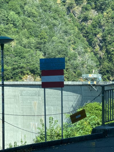 In Gordemo, Switzerland, a concrete dam is visible, a man-made structure of considerable size. The dam's surface is textured and shows signs of weathering, with faint vertical streaks of rust-like discoloration. A chain-link fence runs along the top of the dam, and a series of metal chains are suspended in front of it at varying heights, creating a barrier.
To the left of the dam, a tall, dark lamppost stands, its design utilitarian. Attached to it is a collection of stickers and graffiti, adding a layer of urban art to the scene.
In front of the dam, two prominent signs are displayed on metal posts. The larger sign, positioned centrally, is rectangular and blue, bearing text in white. Above this text, a smaller blue and white emblem is visible. Below the blue sign, a rectangular sign with horizontal stripes of red and white is affixed, representing a national flag.
Further to the right, closer to the viewer and hanging at an angle from the chains, is a smaller, diamond-shaped yellow sign with black text. This sign also appears to convey a warning or prohibition, with text in multiple languages.
The background of the image is dominated by a steep, densely forested mountainside. The trees are lush and green, suggesting a temperate climate. The sunlight casts shadows, indicating it is daytime.
A complex structure, possibly a platform or scaffolding for recreational activities like bungee jumping, is visible on top of the dam in the distance, with some individuals present on it. The overall scene depicts a functional landscape that also incorporates elements of recreation and public information.