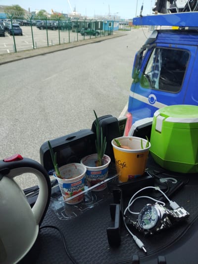 The image shows a close-up view of a dashboard or interior of a vehicle, likely a truck, with a focus on several small potted plants. In the foreground, to the left, is a white electric kettle. To the right of the kettle, on the dashboard, are three small containers with what appear to be sprouting onions. Two of the containers are yogurt cups from the brand "Milssani Alpin Khur". One is a yellow McCafé cup. Behind these pots is a black electronic device, possibly a dashcam or GPS unit. Further to the right, a bright green insulated cooler is visible. In the lower right corner, a silver analog watch with a metal band is connected by a white cable to a power source. The background shows an outdoor scene through the vehicle's window, with a parking lot filled with cars, a fence, and buildings in the distance. The scene appears to be set in a port or industrial area, possibly near the coast. The lighting suggests it is daytime with overcast skies. There are no people visible in the image. The presence of the kettle, plants, and cooler suggests a potentially long journey or a setup for prolonged use within the vehicle. The overall impression is one of a functional vehicle interior with personal touches for the driver. The location is identified as Cuxhaven, Germany, which is a port city on the North Sea coast, known for its maritime activities and industrial areas, consistent with the background imagery.