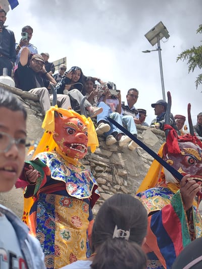 This image captures a vibrant cultural event, likely a religious or festival celebration, in Stingri, India. The foreground features two individuals in elaborate traditional costumes, wearing striking masks. The mask on the left is bright orange with multiple eyes and sharp teeth, adorned with yellow fabric. The costume is richly decorated with colorful embroidered patterns in blue, yellow, red, and green. The second masked figure, on the right, is partially obscured but appears to have a dark red mask with horns and red fabric accents. A black sword or staff is held by one of the masked figures.
Behind the performers, a crowd of onlookers is seated on a stone wall, many of whom are observing the event and using their mobile phones to capture the spectacle. The people in the crowd are dressed in casual clothing. The setting is outdoors, with a cloudy sky overhead. A solar-powered street lamp is visible in the background, suggesting a contemporary setting within a possibly remote or traditional area. The overall atmosphere appears festive and engaging, with a sense of community gathering to witness this cultural performance. The time of day appears to be daytime, given the lighting conditions. There is no visible text in the image.