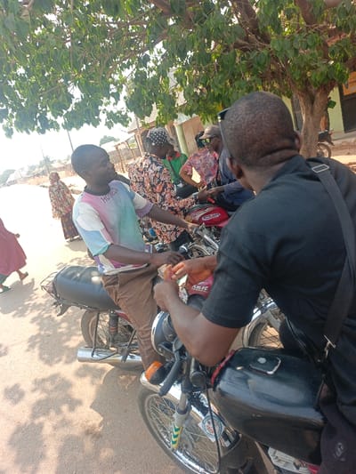 The image displays an outdoor street scene in Nassarawan Eggon, Nigeria, during daylight hours. In the immediate foreground on the right, a male individual with dark, short hair is seated on a black motorcycle, viewed from behind. He wears a black short-sleeved collared shirt and a black shoulder bag. Directly facing him, slightly to the left, another male individual with dark, short hair is seated on a red motorcycle featuring a black seat and a rear metal luggage rack. This individual wears a multi-colored t-shirt with "HORIZON" text and light brown trousers. His hands are extended towards the first individual, holding small, light-colored objects, suggesting a transaction or exchange.
Behind this central interaction, a group of people is visible. To the right of the red motorcycle, a male wearing a dark patterned shirt and a patterned cap is seated on another motorcycle. Next to him, a male wearing a light patterned shirt, sunglasses, and a cap is also seated on a motorcycle. Further left in the background, two individuals are walking away from the central scene: one female in a long patterned garment and another person in a red garment, partially obscured.
The ground surface is unpaved, appearing as dirt or gravel. A large tree with dense green foliage overhangs the upper portion of the scene, providing shade. In the background, several single-story buildings with corrugated metal roofs are visible. One building on the right features a partially legible sign that reads "POST OFFICE."