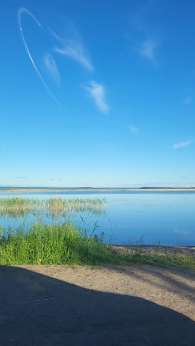 The media file displays a scenic landscape with a vast expanse of clear blue sky on the left, featuring a few wispy clouds and a circular contrail. The right side of the image is dominated by a tranquil body of water, likely a lake or a wide river, reflecting the blue sky and distant, hazy land formations. Along the shoreline, tall reeds and lush green grass are visible, with a dirt path or road bordering the grass on the far right. The sunlight suggests it is daytime, likely late afternoon or early evening given the long shadow cast by an unseen object on the road. The overall mood is serene and peaceful, evoking a sense of calm nature. There is no visible text within the image. The location context of Solnechnoe, Russia, suggests a possibly rural or coastal setting.