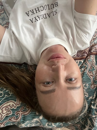 A selfie of a woman lying on a patterned surface, possibly a bed or couch. She has fair skin, light eyes, and light brown hair visible at the top left. She is wearing a white t-shirt with text printed on it. The background features a fabric or surface with a complex pattern of swirls and lines in shades of green, brown, and cream. There is no specific information about Chemoshur, Russia, to determine the location.