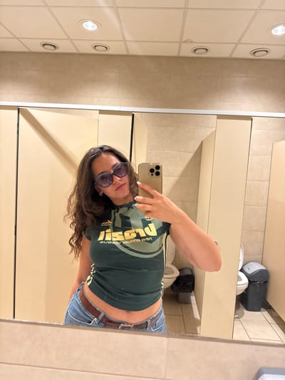 A young woman with long brown hair and sunglasses is taking a selfie in a public restroom. She is wearing a dark green t-shirt and light blue denim jeans. The bathroom has beige walls, a white tiled ceiling with recessed lighting, light beige stall doors, a toilet, and a toilet paper dispenser. The location is unknown.