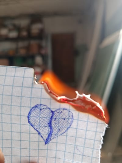 A close-up shot captures a piece of graph paper with a blue ink drawing of a heart. The top edge of the paper is burning, with flames and smoke rising. The background is out of focus, showing shelves with various objects, possibly in a room or workshop. The lighting suggests it's daytime, with sunlight streaming in from the right, creating a bright glare. The image does not provide any specific information or context related to Sokuluk, Kyrgyzstan.