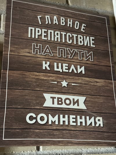 The image shows a sign with a wood-grain background mounted on a brick wall. The sign reads, "The main obstacle on the way to the goal: Your doubts." The text is in white Cyrillic letters. The location is Zamankul, Russia. There are no people present.