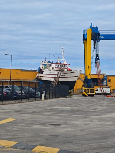 This media file depicts a ship in a dry dock with a large industrial crane nearby. The ship is white with a red stripe and the number "13 657" is visible on its side. A wooden gangway leads from the ground to the ship. The industrial crane is a prominent feature, painted in yellow and blue, with a "350t" label. Several cars are parked behind a fence in the foreground, and a white van is visible near the crane. A long yellow building serves as a backdrop. The scene appears to be in an industrial or port area, possibly a shipyard. The sky is cloudy, suggesting overcast weather. There are no people visible in the frame. The overall atmosphere is industrial and operational. The location is identified as Boulogne-sur-Mer, France, which is consistent with a coastal port setting. The image captures a moment of stillness, with the ship out of the water, likely undergoing maintenance or repair.