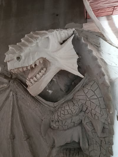 This media file captures a close-up, side-view of a white, unfinished sculpture of a dragon's head and upper body, likely made of plaster or cement. The dragon has an open mouth revealing sharp, pointed teeth and a detailed tongue. Its head is adorned with jagged crests, and its body appears to be covered in scales, with some sections intricately carved to resemble stone mosaic work. Partially visible are large, folded wings with a textured, leathery appearance. The sculpture is mounted on a rough, grey wall, with another wall made of pinkish material visible in the upper right corner. The lighting suggests an indoor setting, possibly a workshop or an area under construction. There is no visible text. The scene is static, focusing on the artistic creation. The context provided indicates the location as Smarhon, Belarus.