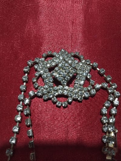 The image, captured in Domažlice, Czechia, depicts a close-up, overhead view of a vintage-style piece of rhinestone jewelry. The central subject is an intricate, silver-toned metal medallion heavily encrusted with numerous small, clear, faceted rhinestones. The medallion features a prominent diamond-shaped cluster of stones at its core, surrounded by an ornate, openwork design composed of loops and curves, also densely paved with rhinestones. Extending from both sides of this central medallion are multiple strands of rhinestones, forming flexible chains that drape downwards and out of the lower portion of the frame. Each chain consists of individual clear rhinestones set in silver-toned metal, linked linearly. The jewelry item rests on a deep red, textured fabric background, which exhibits a slight sheen and subtle folds. The lighting appears direct, causing the rhinestones to sparkle and highlighting the texture of the fabric. No individuals, actions, or other objects are present in the scene.