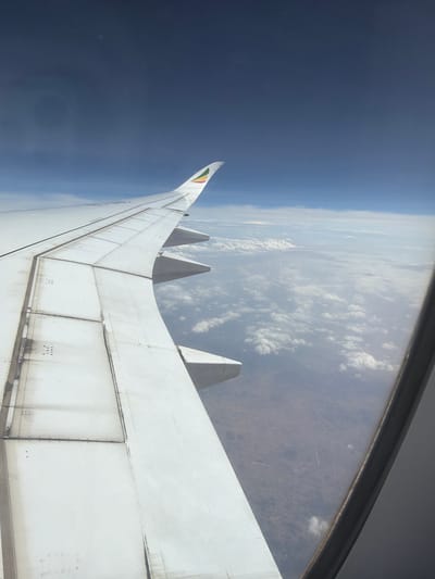 The image presents an aerial view from an airplane window, looking along the aircraft's wing towards the sky and ground below. The perspective is tilted, showing the horizon line as almost vertical.

The prominent subject is the clean white airplane wing, extending from the lower right of the frame towards the upper left. The wing's surface displays distinct panel lines, rivets, and areas of grime and dirt, especially on the lower sections. Several trapezoidal fin-like structures, likely vortex generators or wing fences, are visible on the upper surface of the wing. The wingtip curves upwards into a winglet, adorned with a colorful emblem in green, yellow, and red. These colors are indicative of the Ethiopian flag, strongly suggesting the aircraft belongs to Ethiopian Airlines and the flight is either departing from, arriving at, or flying over Ethiopia, consistent with the Addis Ababa location context.

The sky transitions from a dark, deep blue on the far left, implying the high altitude, to a lighter blue on the right, where scattered white cumulus clouds are visible. Through and beneath these clouds, a brownish, earthy landscape with subtle topographical details and faint lines, possibly indicating natural features or human infrastructure, can be seen. The view is framed by the dark gray or black outline of the airplane window along the top and right edges.

The scene depicts a flight during daytime, with clear visibility. The weather appears partly cloudy over the land, with a clear upper atmosphere. No specific text is discernible within the image.