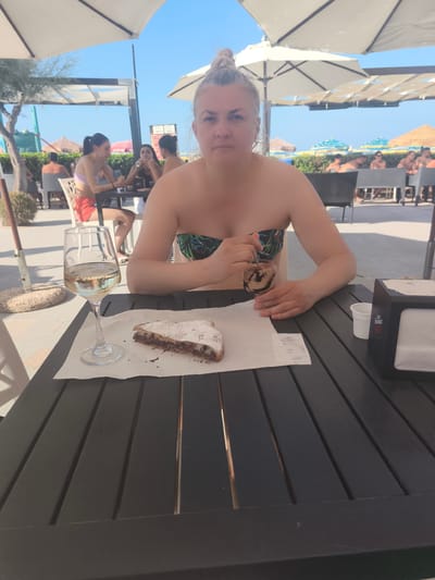 The image depicts a woman sitting at a dark wooden table outdoors, in what appears to be a cafe or restaurant setting at a beach resort. She is facing the camera and has her hair pulled up in a bun. She is wearing a strapless bikini top with a tropical leaf pattern. In front of her on the table is a glass of white wine, a slice of pastry dusted with powdered sugar, and a receipt. She is holding a spoon in a small glass, possibly for stirring a drink or eating dessert. Other people and beach umbrellas are visible in the background, suggesting a sunny day at the beach. The general atmosphere is one of relaxation and leisure. While the specific location in San Salvo, Italy is not explicitly stated in the image, the context suggests a coastal establishment.