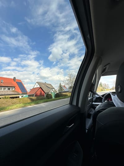 The image shows a view from inside a car, looking out the passenger-side window. The sky is visible, with scattered white clouds against a blue background. Outside the window, houses line the street. The houses have red tiled roofs. Solar panels are visible on one roof. The location is Gunzenhausen, Germany, based on visual elements like architectural style common in the region. The car interior, including the dashboard and seating, is partially visible, as well as the driver, with a partially obscured face.