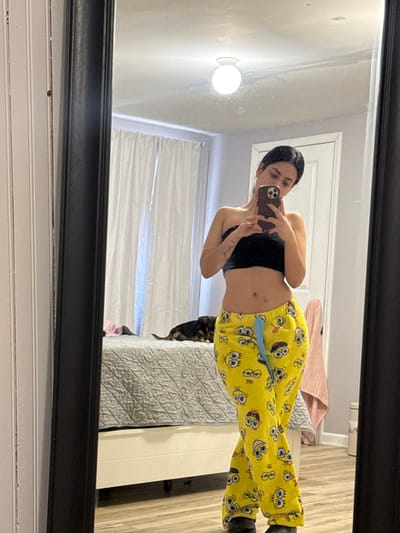 A woman takes a mirror selfie wearing a black top and yellow SpongeBob pajama pants. She holds her phone in her right hand. A dog is partially visible near the bed. The room contains white walls, a white headboard, a gray quilted bedspread, and white curtains. A light fixture is on the wall near the mirror. The mirror has a black frame.