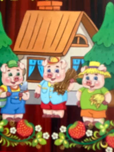 The image depicts a cartoon scene of three pigs in front of a house. The pig on the left wears a green cap, yellow shirt, and blue overalls, holding a shovel and a tool. The middle pig is dressed in a blue shirt and red shorts, holding a bundle of straw. The pig on the right wears a yellow hat with a purple band, a green shirt, and orange shorts, carrying a small bundle of straw. The house behind them has an orange roof and white walls with blue curtains in the windows. Two green cartoon trees flank the house, and the foreground features a decorative border with strawberries and clover leaves. There is no discernible information about the city of Edem, Russia in this image.