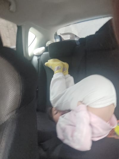 A child is seen sleeping or resting comfortably in the back seat of what appears to be a car. The image is rotated, showing the child curled up on their left side, facing towards the left side of the frame. The child is wearing a light-colored, possibly tie-dye pink, hooded sweatshirt and white pants. Their feet are adorned with bright yellow socks that feature a repeating pattern, possibly of cartoon characters or unicorns.

The child's face is partially visible and appears peaceful, suggesting they are in a deep sleep or relaxed state. They are lying across the dark fabric of the car's back seat. Above the child, part of a car seat's headrest or backrest with a textured gray fabric is visible. To the right of the child, the interior structure of the car, including a window, is visible, indicating daylight from outside. The overall setting is the interior of a vehicle, possibly during a journey, with the child taking a nap. No text is visible in the image.