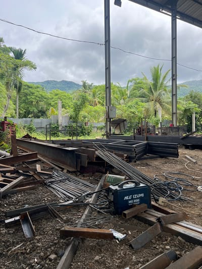 The image depicts an outdoor scene at a construction or fabrication site, possibly in Lambarobiluy, Indonesia. The foreground is filled with a disorganized collection of metal construction materials, including large I-beams, rebar, and various steel shapes, lying on the ground amidst dirt and gravel. A blue NLG IZUMI MO 200 welding machine is visible in the mid-foreground, with coiled black cables nearby. In the background, a corrugated metal fence separates the site from a lush, green environment of trees, palm trees, and rolling hills under a cloudy sky. Several industrial metal support columns are present, suggesting an open-air structure. There are no people visible in the image.