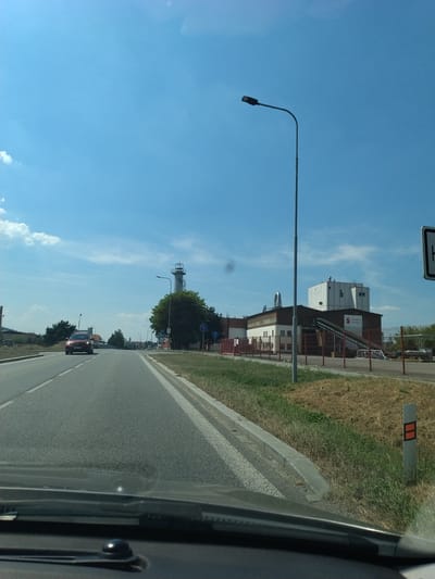 This is a daytime, eye-level, outdoor shot from a moving vehicle, looking forward down a paved road in Horažďovice, Czechia. The sky is bright blue with scattered white clouds. On the right side of the road, there is a large industrial building complex with white and brown sections. A conveyor belt system is visible leading from a lower structure to a taller building, which has the text "Lyckeby Arrowflex" on it. A tall, grey, cylindrical structure resembling a tower with a spiral staircase is visible in the distance on the right, behind a cluster of trees. A red car is traveling in the same direction as the camera vehicle, in the left lane. A street lamp stands on a pole on the right side of the road, near the industrial complex. Several traffic signs and fences are also present along the road and around the industrial site. The foreground shows the dashboard of the vehicle.