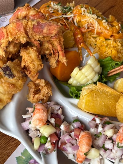 The image shows a close-up of a seafood platter served on a white divided plate. The platter contains several distinct dishes. In the upper left quadrant, there is a pile of deep-fried seafood, appearing to be shrimp or langoustines, with a reddish hue visible through the golden-brown batter. To the right of the fried seafood is a mound of yellow rice, topped with more langoustine tails and a creamy orange sauce, garnished with finely chopped herbs and shredded white cheese. Adjacent to the rice is a piece of boiled corn on the cob, resting on a bed of green lettuce. In the lower section of the plate, a colorful ceviche-like salad is presented in the curved sections of the plate. This salad contains diced red onion, tomato, white onion, cilantro, and pieces of shrimp or langoustine, with some green peas and a light-colored dressing. There are also a few pieces of fried plantains or yuca in the lower right quadrant. The background shows a wooden table surface. The establishment appears to be a restaurant in San Vicente de Azpitia, Peru, known for its seafood offerings.