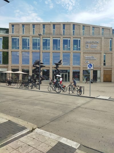 In Monheim am Rhein, Germany, a modern building with a light-colored brick facade stands under a partly cloudy sky. The building features a repeated pattern of large, rectangular windows, with some sections of greenery visible on the left side. Signage on the building indicates "EMOTION center Gusek," as well as "WOOLWORTH" and "DEICHMANN." A blue and white sign depicting people and bicycles signifies a "ZONE."
In the foreground, a paved area stretches out, with a section of tiled walkway on the left leading to a curb. Several bicycles are parked or in use on the paved area. A woman is seen walking a bicycle on the left. In the middle, a group of people are on bicycles, some are riding, and others are stationary. One man is riding a bicycle towards the right. In the background, through the glass windows of the building, merchandise and displays are visible. Two abstract, dark sculptures are positioned in front of the building's entrance. Several outdoor tables and umbrellas are set up on a patio area to the left of the entrance. The overall scene suggests a public space, likely a pedestrian zone or plaza in front of a commercial building.