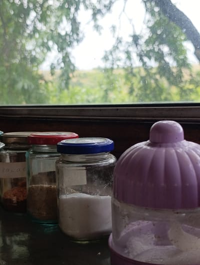 A close-up, slightly tilted, eye-level shot shows several glass jars with various contents lined up on a dark, reflective surface. Behind the jars, a window with a wooden frame reveals a blurred view of green foliage and a hint of a landscape under an overcast sky. The first jar on the left has a metallic lid and a handwritten label that appears to read "kakao". Inside, there is a reddish-brown granular substance. The next jar has a red lid and contains a light brown powder. The third jar from the left has a blue lid and is filled with a white granular substance, possibly sugar or salt. To the right of these jars, and dominating the foreground, is a portion of a purple plastic container with a ribbed, dome-shaped lid, also containing a white substance. The overall impression is one of a simple, perhaps rustic, storage area, potentially in a kitchen or pantry. The scene is set in the village of Mladinovo, Bulgaria.