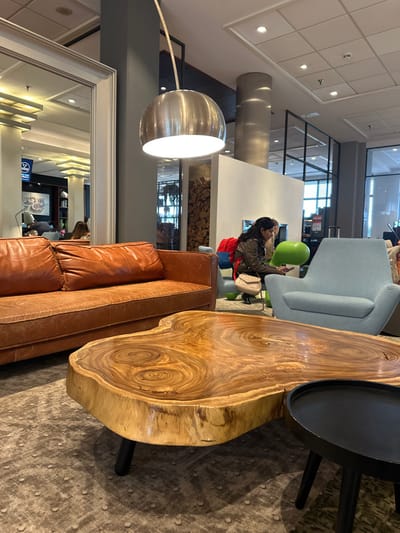 The image depicts an indoor lounge area, identified as a Mercure hotel lobby in Hoofddorp, Netherlands, via a reflected 'MERCURE' sign in a mirror. The foreground features a large, irregularly shaped, live-edge natural wood coffee table with black legs, positioned on a grey patterned carpet. Adjacent to it is a smaller, round black coffee table. To the left, a tan leather sofa is partially visible. To the right, a light blue fabric armchair is present, with a bright green sculptural chair situated behind it.

In the mid-ground, a female individual with dark hair is seated, wearing a dark jacket and light-colored pants, looking down at a mobile device held in her hands. A light-colored shoulder bag rests on her lap, and a red backpack is on the floor to her immediate left. Overhead, a large, illuminated silver spherical pendant lamp hangs from the ceiling. Behind the seated individual is a white partition wall, beyond which stacked firewood is visible, along with glimpses of glass panels or windows and additional seating. A large, white-framed wall mirror on the far left reflects other areas of the interior and the aforementioned 'MERCURE' branding.