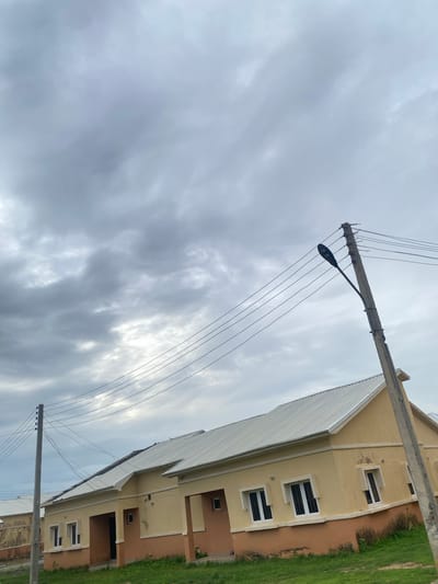 A low-angle, vertical, full shot captures a street scene under a heavy, overcast sky, with light filtering through broken clouds. In the foreground, a concrete utility pole with a street lamp attached stands on the right, supporting multiple electrical wires that crisscross the frame. To the left, another, thinner utility pole with insulators is visible. The middle ground features a row of tan and light orange single-story houses with white roofs. The houses have dark-framed windows and appear to be under construction or newly built, with some visible staining on the lower exterior walls. A patch of green grass is in front of the houses. The sky dominates the upper portion of the image with its dense, gray cloud cover, hinting at approaching rain.