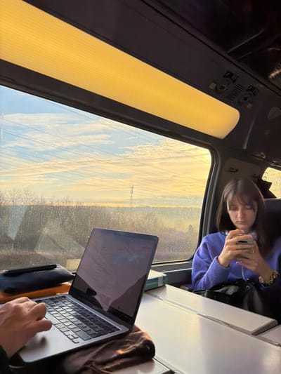 This image captures an interior view from a train car, located in Soignolles-en-Brie, France. Golden sunlight illuminates the scene, suggesting either dawn or dusk.

In the left foreground, the hands of one person are visible, operating a silver laptop resting on a pull-down tray table. A dark zippered case is positioned to the left of the laptop.

To the right, a female passenger with dark hair and bangs occupies a seat, facing forward. She wears a lavender knitted sweater and is engaged with a smartphone held in both hands. A dark bag rests on the seat beside her, and a small green and white object is on the adjacent tray table.

The train interior features ribbed overhead lighting emitting a yellowish glow and control panels, including seat number '891,' above the window. The window glass exhibits condensation or moisture.

The exterior landscape, seen through the window, comprises bare deciduous trees along the horizon, multiple power lines traversing the sky, and a distant cellular tower. The sky displays scattered clouds against a backdrop of blue and warm golden hues.