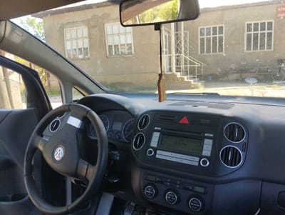 The image shows the interior of a car, looking out through the windshield. The dashboard and steering wheel are visible, along with the car's radio and air vents. Outside the car, a building with several windows and a staircase is visible. There is also a bicycle parked in front of the building. The scene appears to be in a rural or suburban area. A wooden car air freshener is hanging from the rearview mirror. The location is Tătărești, Moldova.